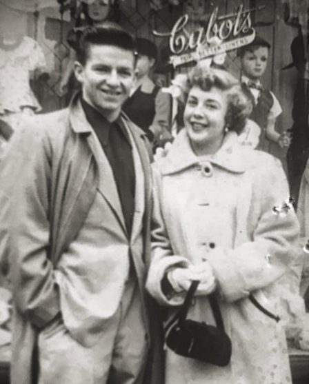 Black and white historical photograph of Mama and Papa D, a smiling Italian couple in New York. Papa D is wearing a dark jacket over a light shirt, and Mama D is wearing a light-colored buttoned coat, with a crowd and a sign reading 'Cabot's' in the background.